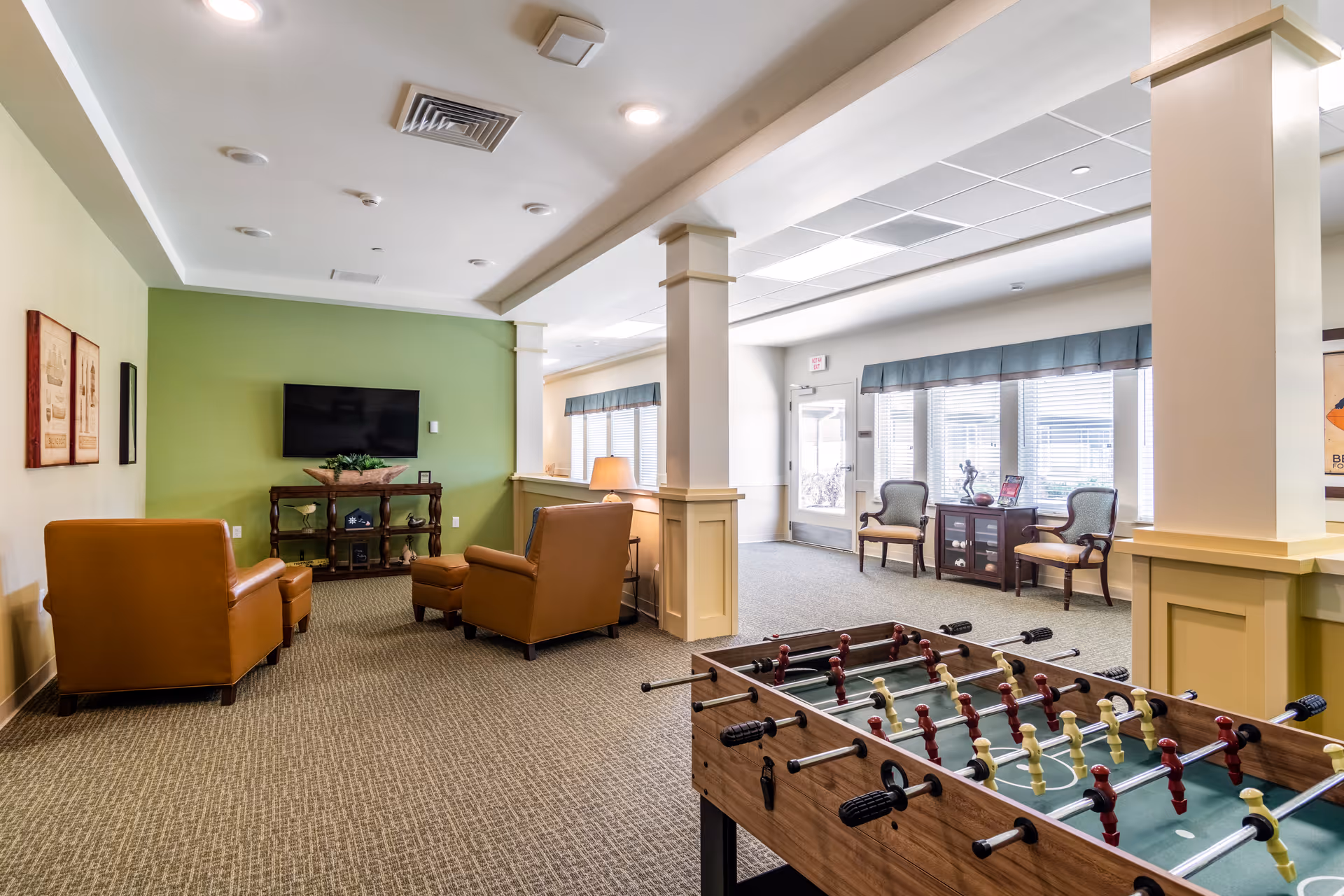 A spacious common area in a senior living facility featuring a foosball table in the foreground, two brown leather armchairs facing a wall-mounted flat screen TV, and a small wooden table with decorative items. The room has green and beige walls, carpeted flooring, and large windows with blinds letting in natural light. There are also two chairs and a small cabinet near the windows.