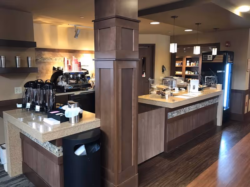 Interior view of a coffee and snack bar area with a coffee machine, stacks of red cups, condiment holders, a trash bin, and a display case with baked goods. The area has wooden cabinetry, a tiled backsplash, and pendant lighting.