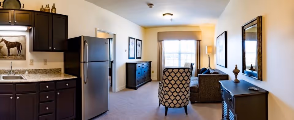 Interior view of a senior living facility room at Magnolia Springs Florence showing a kitchenette with dark cabinets and a stainless steel refrigerator on the left, and a living area with a patterned armchair, sofa, dresser, and window with curtains in the background.