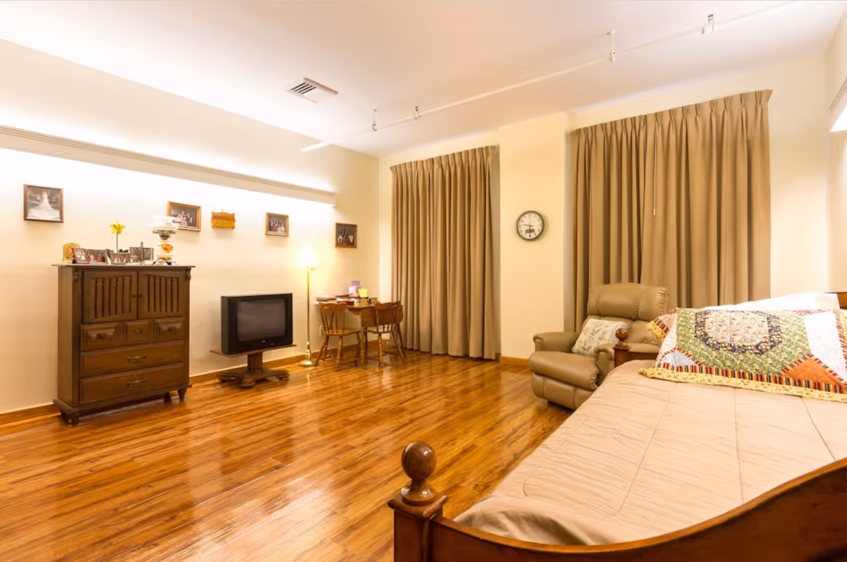 Well-lit assisted living bedroom with wooden floors, a bed with a colorful quilt, a recliner, TV, dresser, and a small dining table.