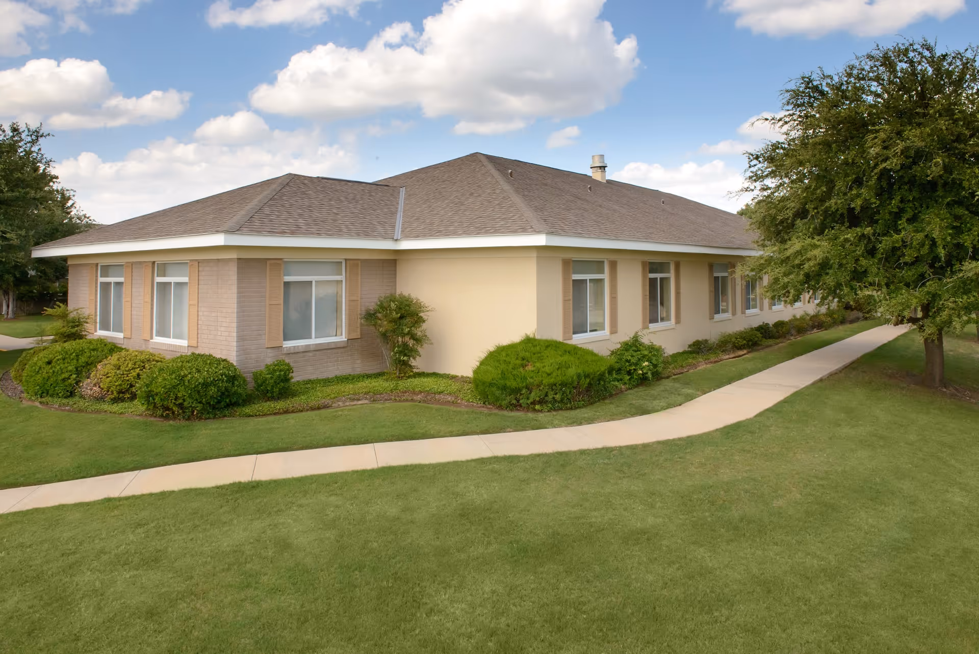 Exterior view of a single-story building with beige walls and a brown shingled roof, surrounded by green grass, bushes, and trees under a partly cloudy sky.