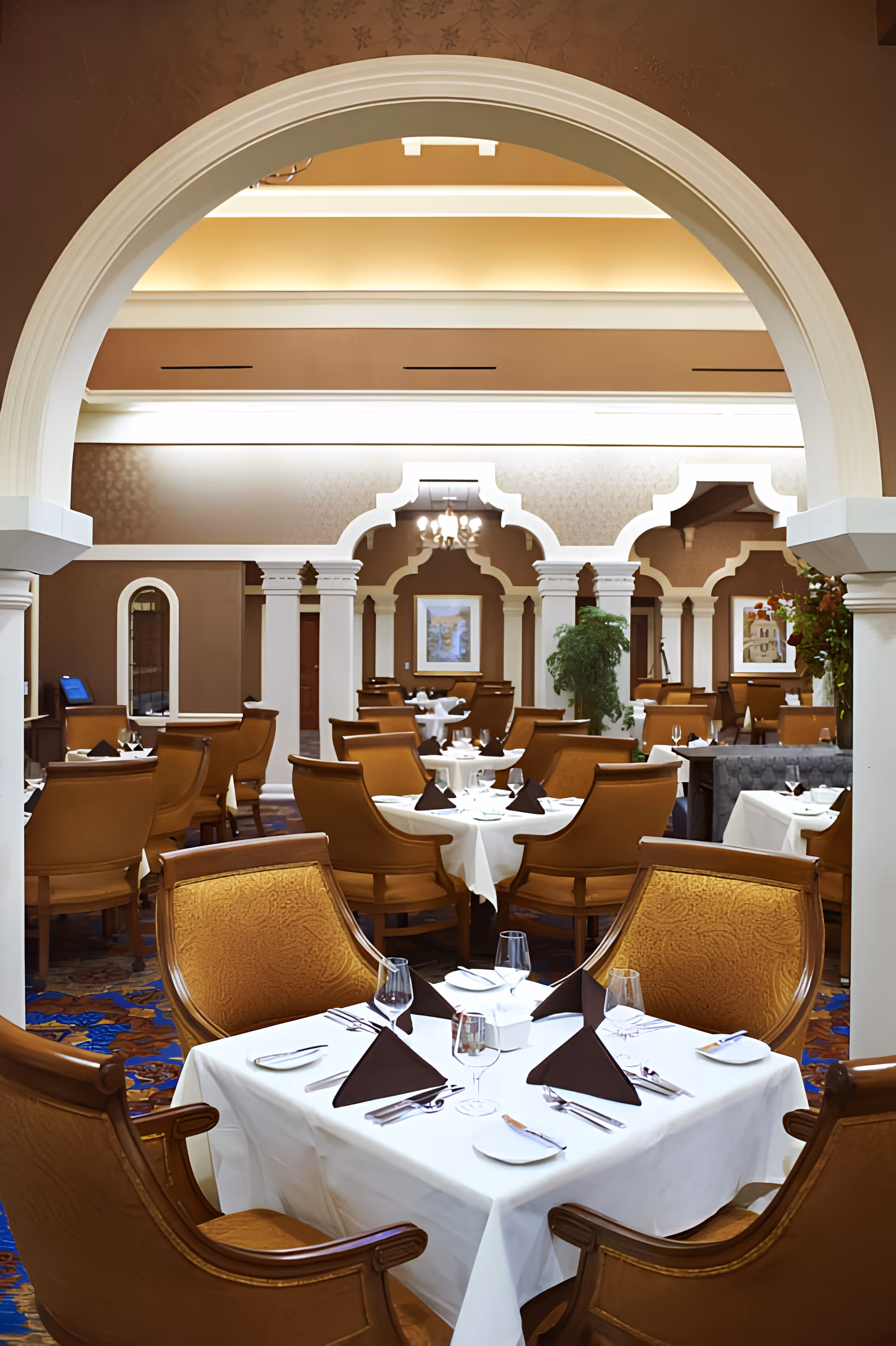 Formal dining room with neatly set white-tablecloth tables and brown upholstered chairs beneath decorative arches.