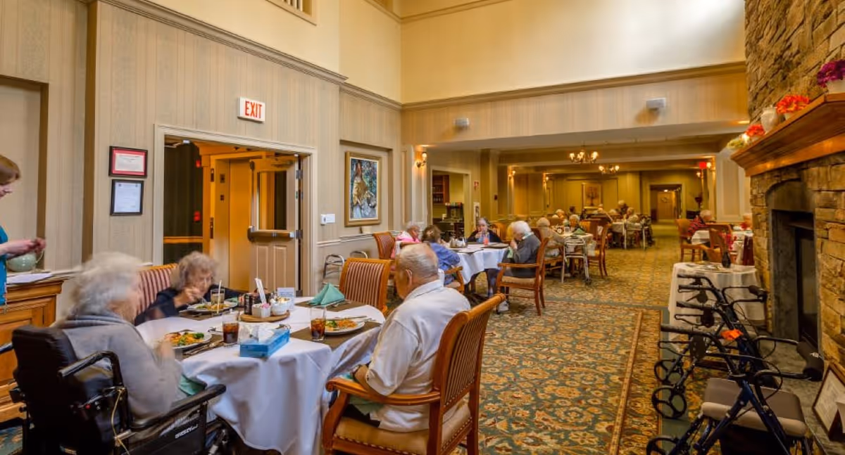 A dining room in a senior living facility with elderly residents seated at round tables covered with white tablecloths, eating meals and socializing. The room features patterned carpet, warm lighting, a stone fireplace on the right, and several walkers near the fireplace. A staff member is standing near one table, and an exit sign is visible above a doorway.