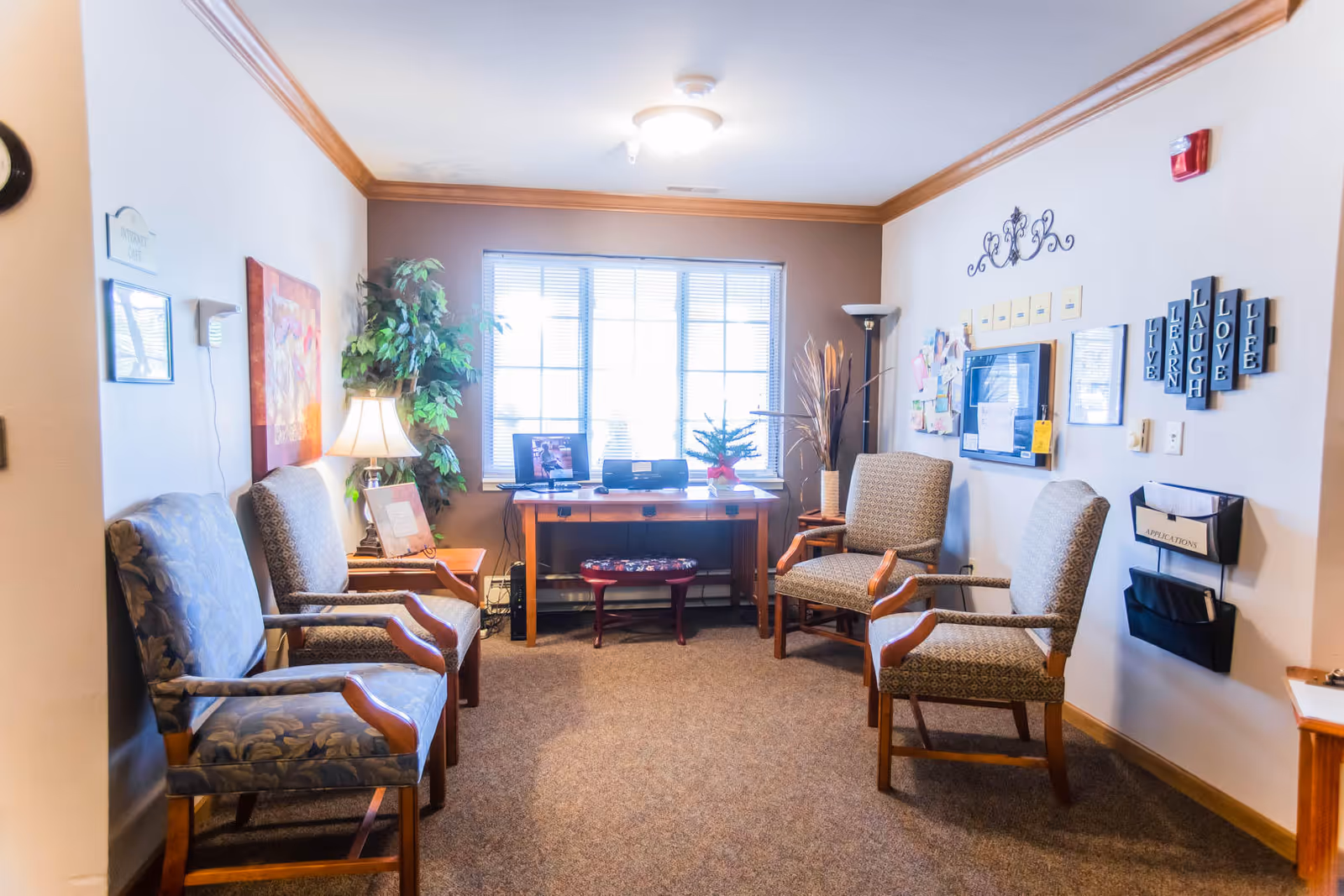 A cozy waiting or common area in Brenwood Park Assisted Living with four upholstered armchairs arranged around a small wooden table. A desk with a computer and printer is positioned in front of a large window letting in natural light. The walls are decorated with framed pictures, a clock, a bulletin board, and inspirational word blocks that read 'LIVE LEARN LAUGH LOVE LIFE'. There are also plants and a lamp adding warmth to the room.