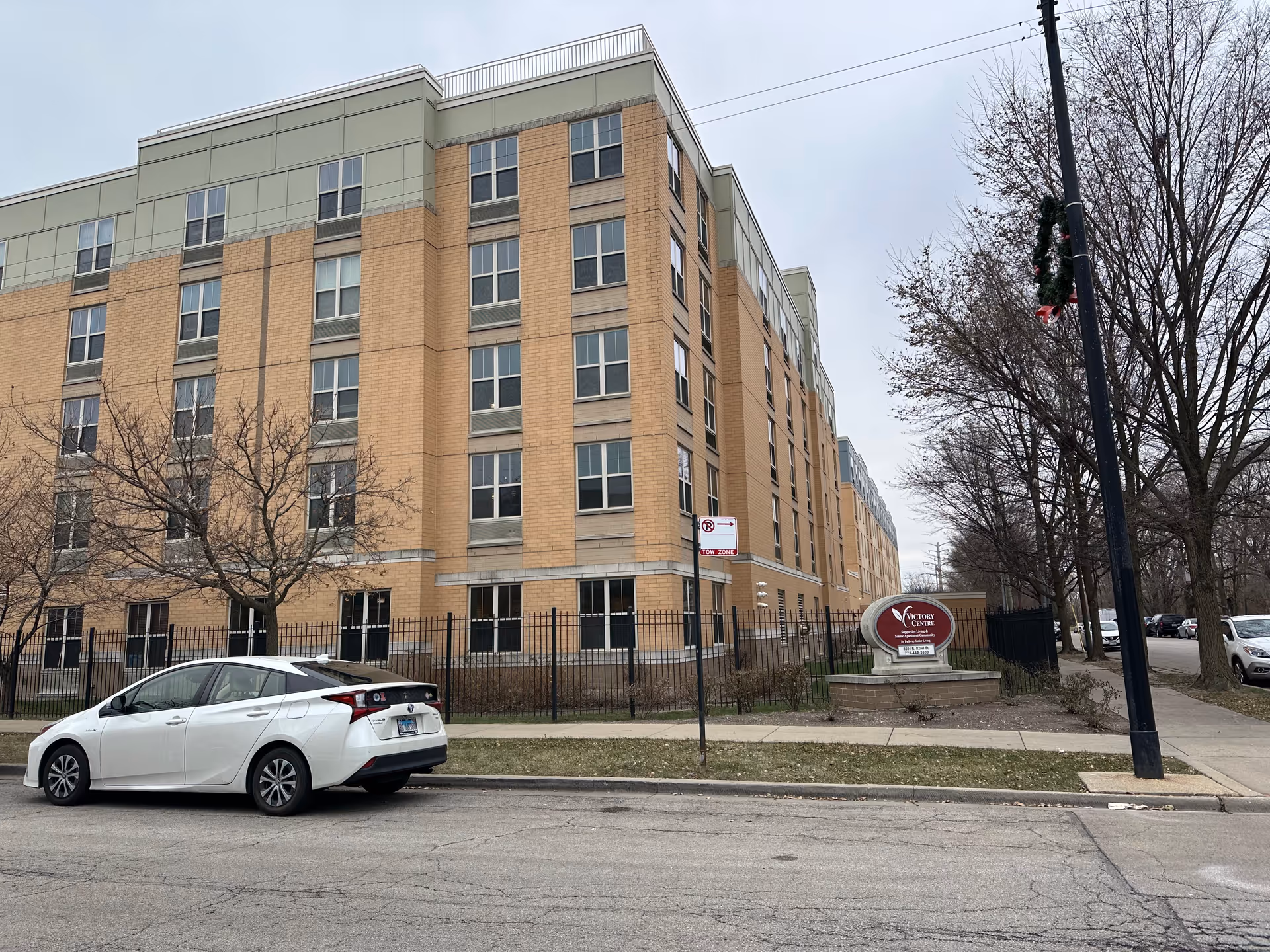 Exterior view of a multi-story brick building with many windows, identified as Victory Centre of South Chicago Supportive Living by a sign near the sidewalk. A white car is parked on the street in front of the building, and leafless trees line the sidewalk under an overcast sky.