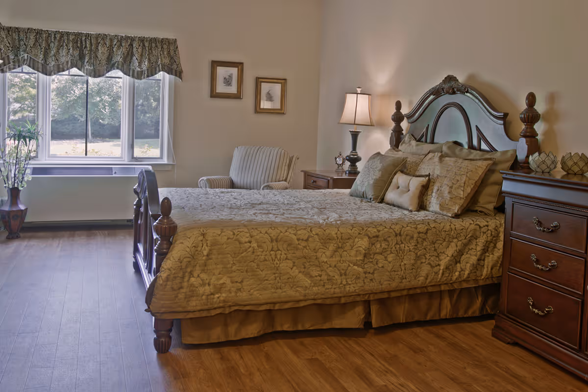 A cozy bedroom with a wooden bed frame and a patterned beige bedspread. The room features a wooden nightstand with a lamp and decorative items, a striped armchair, two framed pictures on the wall, and a large window with a valance overlooking greenery outside. The floor is wooden.