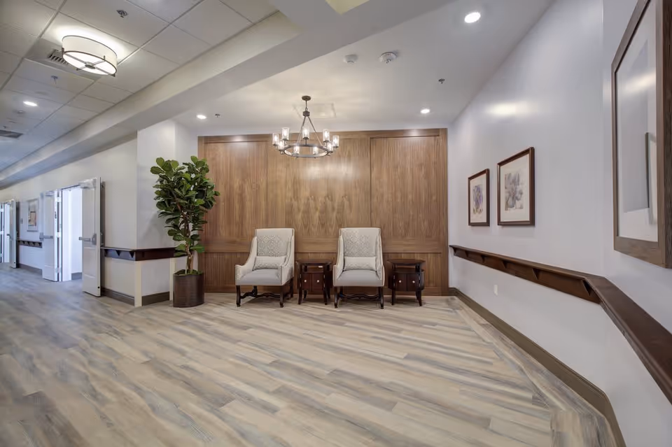 A spacious hallway in a senior living facility with light wood flooring, white walls, and a wooden accent wall. Two upholstered armchairs with patterned cushions are placed against the wooden wall, flanked by two small wooden side tables. A large potted plant is positioned to the left of the chairs. The hallway has ceiling lights, a modern chandelier above the chairs, framed artwork on the walls, and handrails along the walls.