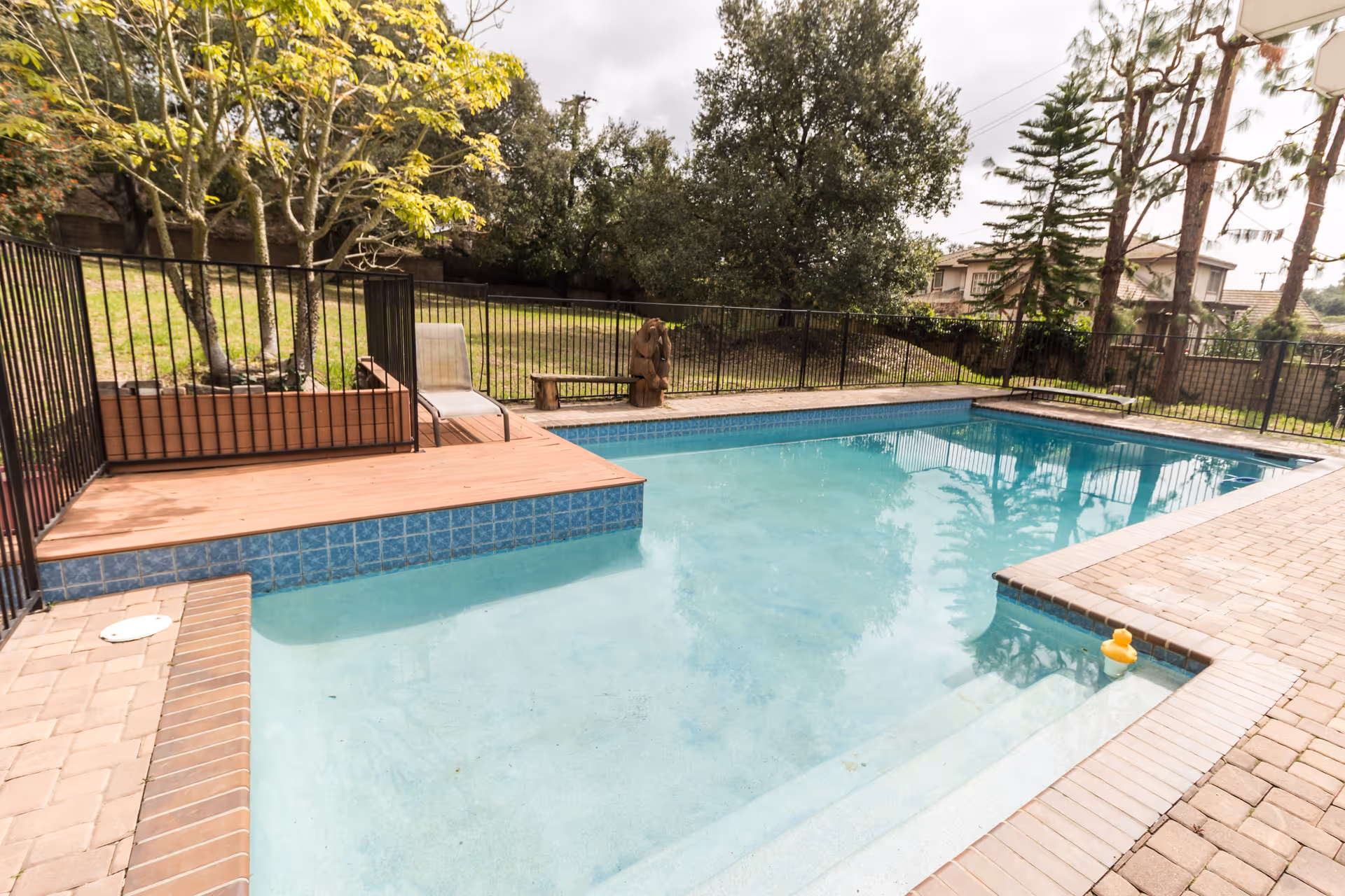 Outdoor swimming pool with clear blue water surrounded by a brick and tile deck, a black metal fence, trees, and a grassy area with a lounge chair and a wooden bench.