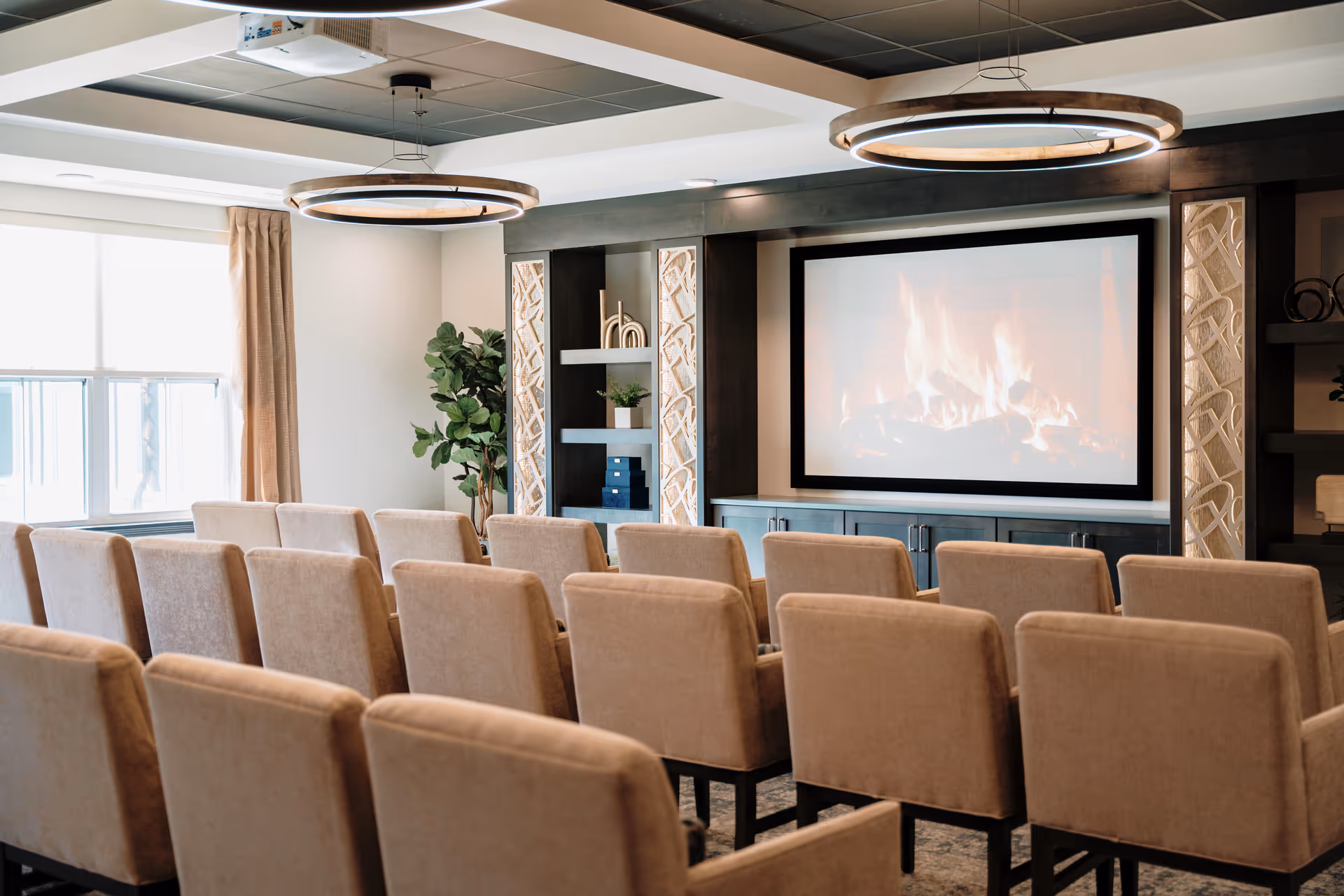 Rows of upholstered chairs facing a large screen showing a fireplace in a cozy community media room.