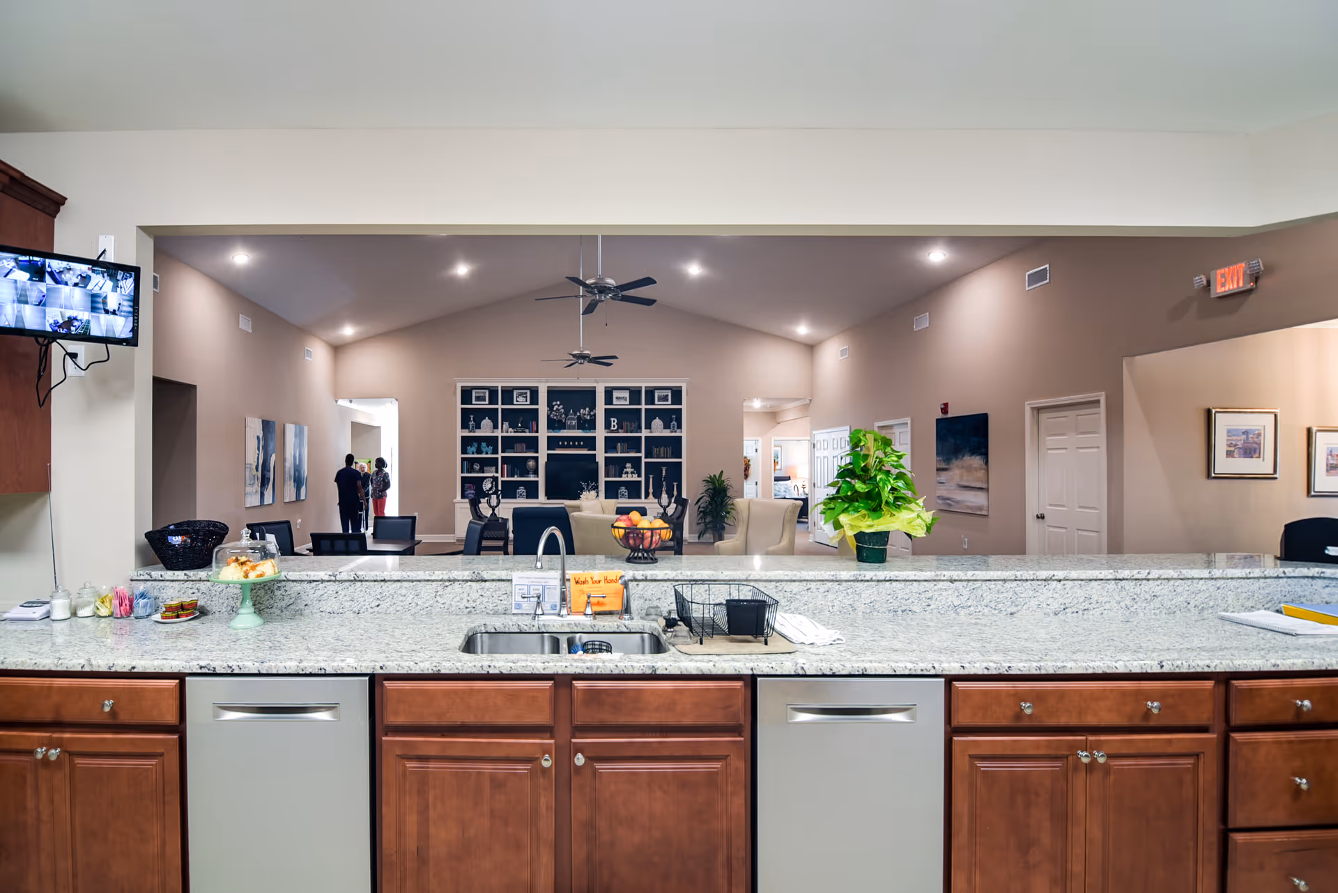View from a kitchen area with granite countertops and wooden cabinets looking out into a spacious living room with beige walls, ceiling fans, built-in shelves, and seating. A small group of people is visible in the background near the entrance. A TV monitor displaying security camera footage is mounted on the left wall.