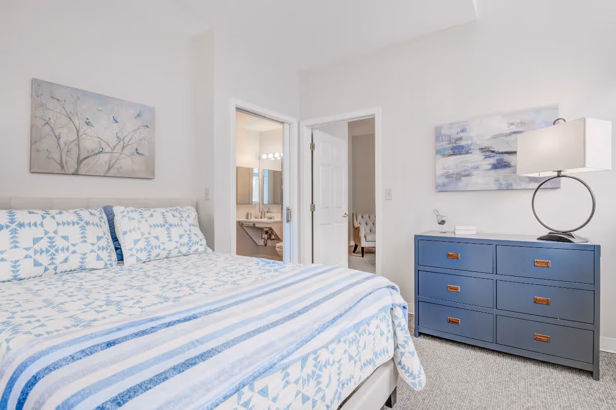 A bright bedroom with a bed covered in a blue and white patterned quilt and matching pillows. On the right side, there is a blue dresser with six drawers, a modern lamp, and decorative items on top. Two open doors reveal a bathroom with a sink and mirror, and another room with a tufted chair. The walls are white, and there are two pieces of artwork hanging above the bed and dresser.