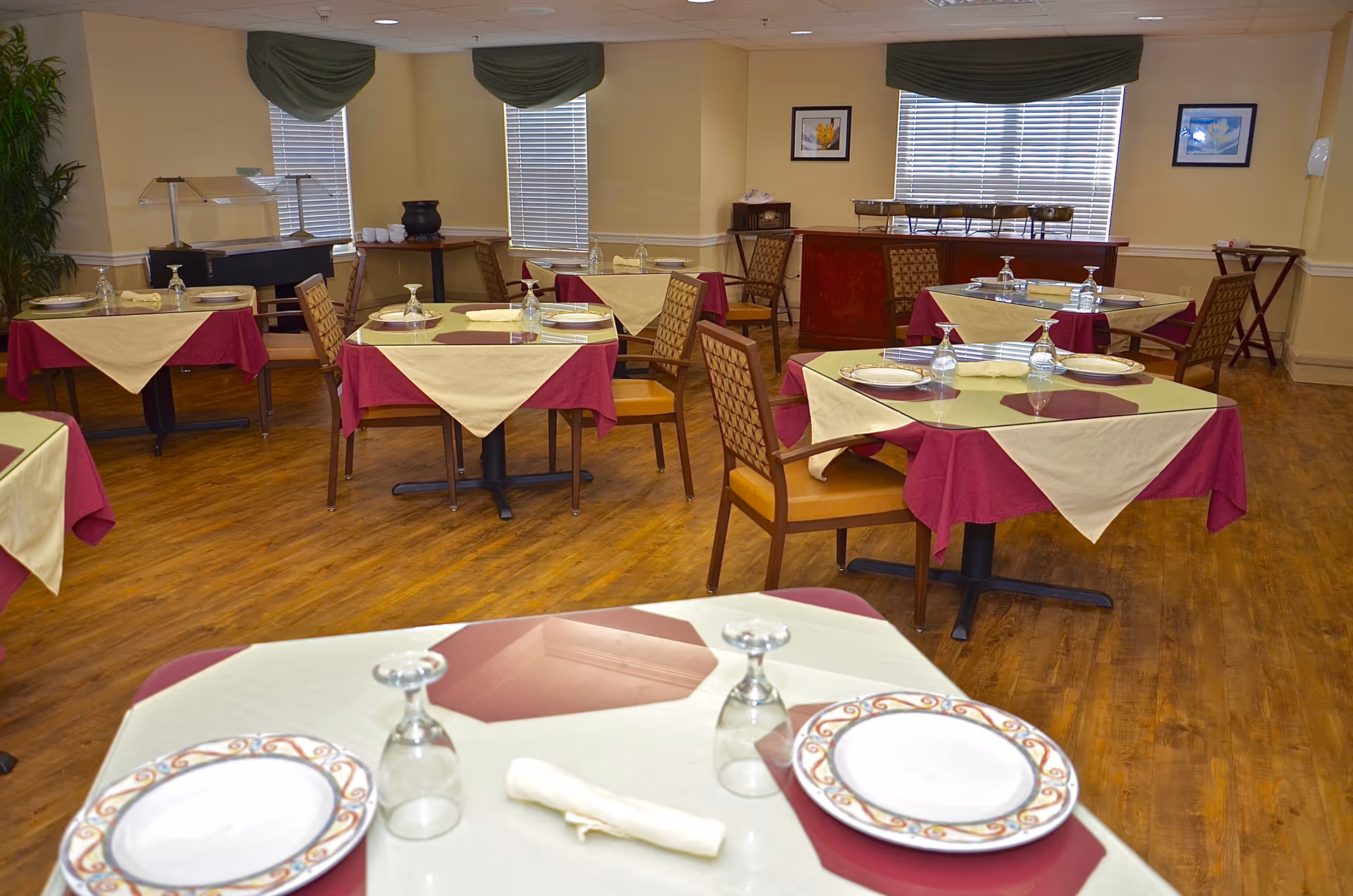 A dining room with several tables covered in burgundy and beige tablecloths, each set with plates, napkins, and upside-down glasses. The room has wooden flooring, windows with green valances, framed pictures on the walls, and buffet tables in the background.