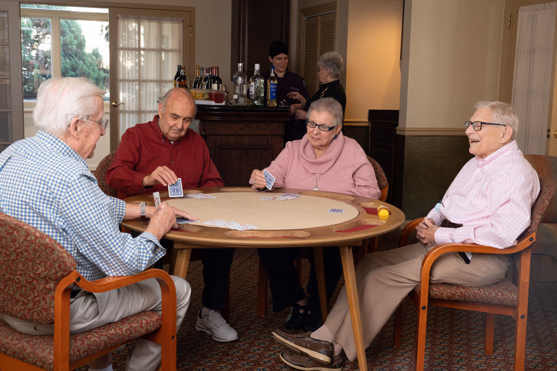 Four elderly people sitting around a round table playing cards in a cozy room with a bar in the background where two women are standing and talking.