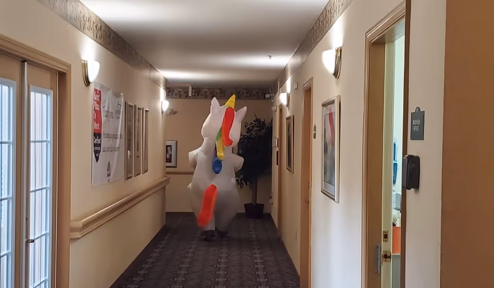 A long indoor hallway with beige walls and carpeted floor, lit by wall sconces. On the right side, there are doors and framed pictures on the walls. At the end of the hallway, a person is walking away wearing a large inflatable unicorn costume with a colorful mane and tail.