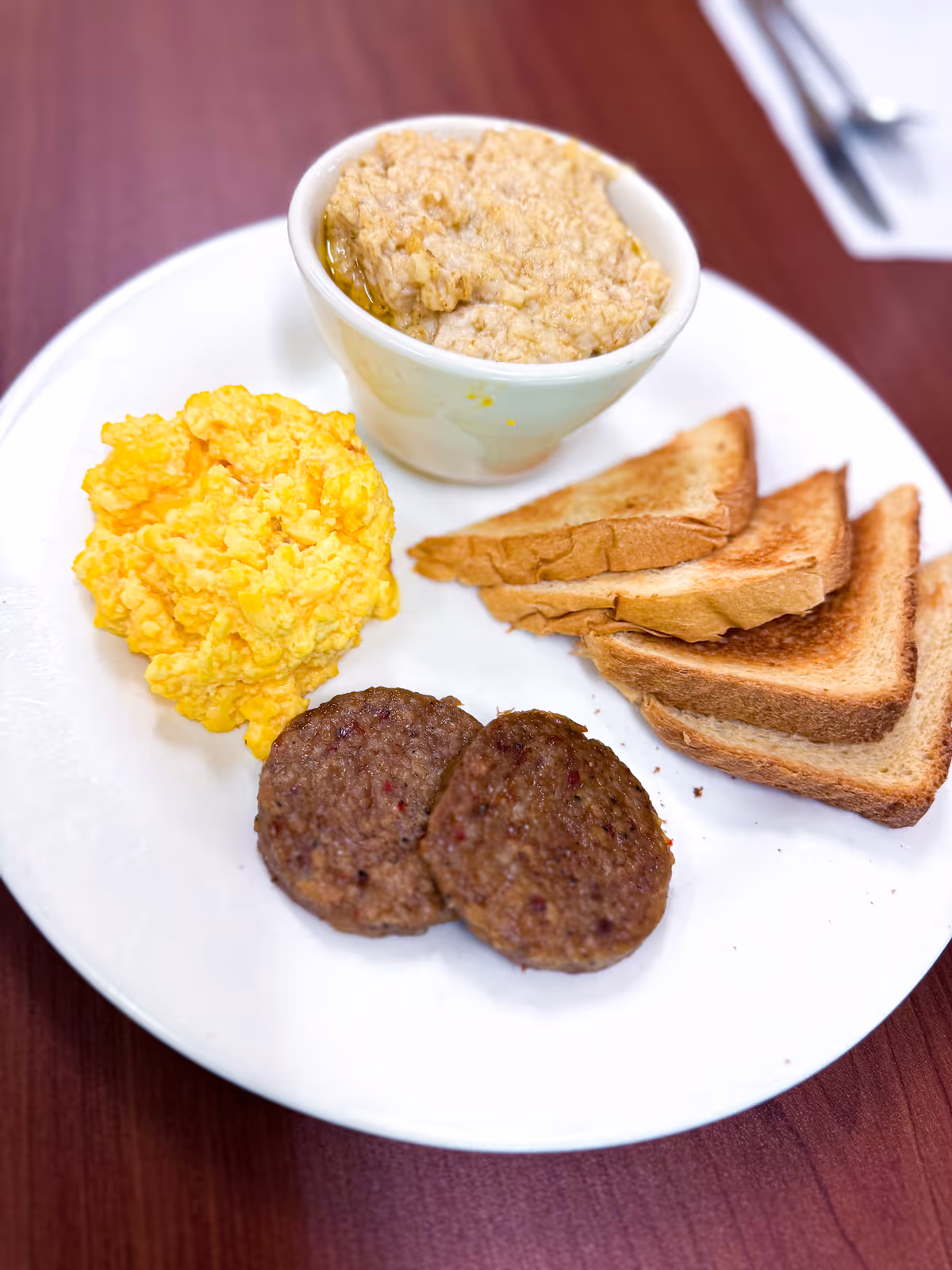 Plate of breakfast with scrambled eggs, two sausage patties, several slices of toast, and a bowl of oatmeal on a table.