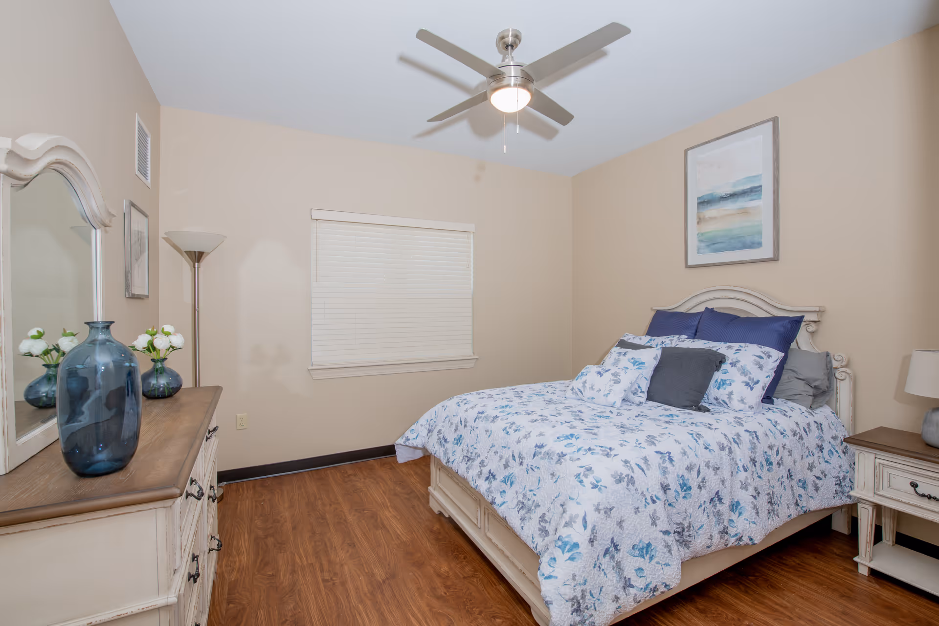 A cozy bedroom with a floral-patterned bedspread on a white wooden bed frame. The room features a ceiling fan with a light, a window with closed blinds, a white dresser with a large mirror and decorative vases, a floor lamp, and a nightstand with a lamp. The walls are painted beige and the floor has wood-style flooring.