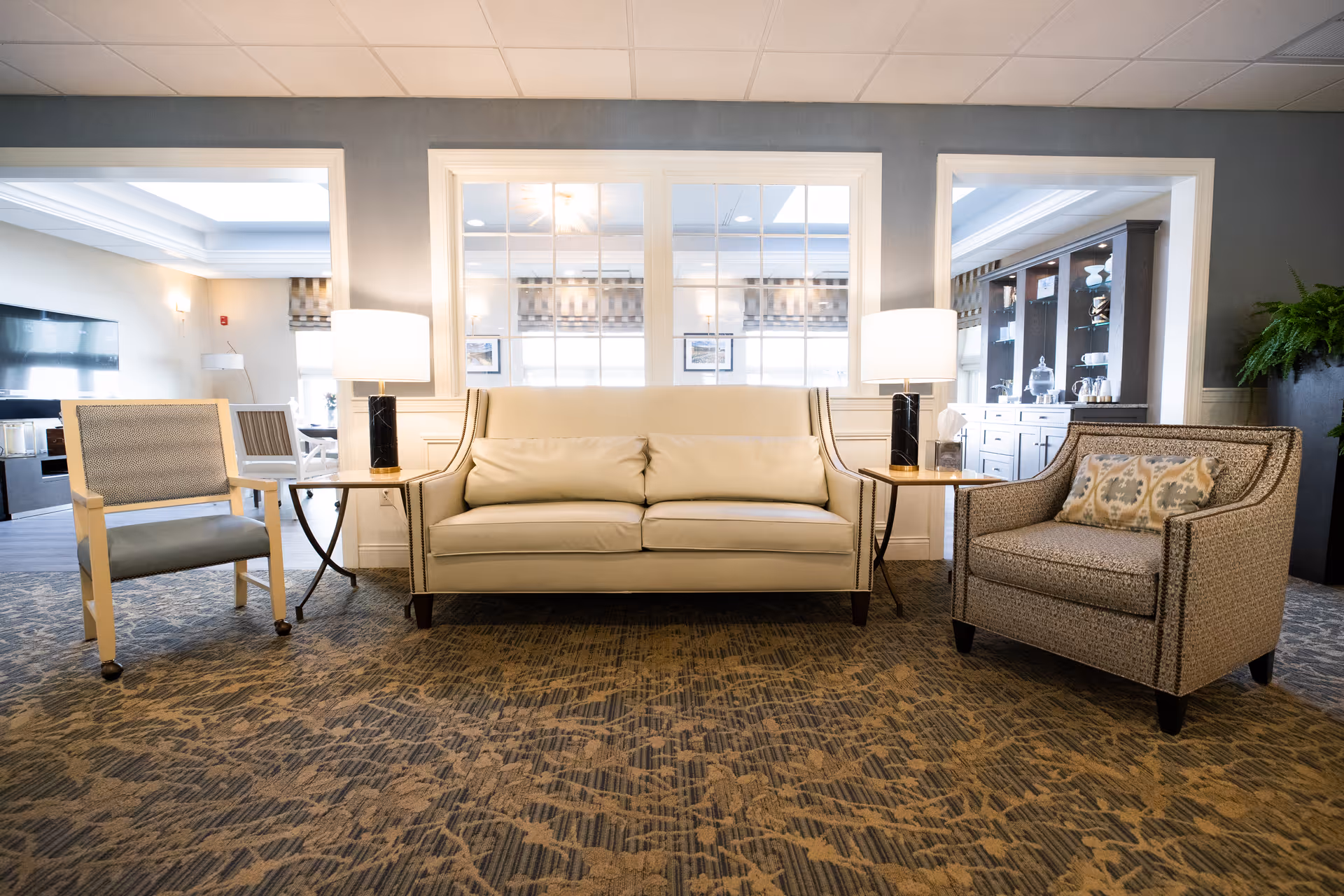 Bright communal lounge with a cream sofa flanked by two armchairs and side tables with lamps in front of interior windows.