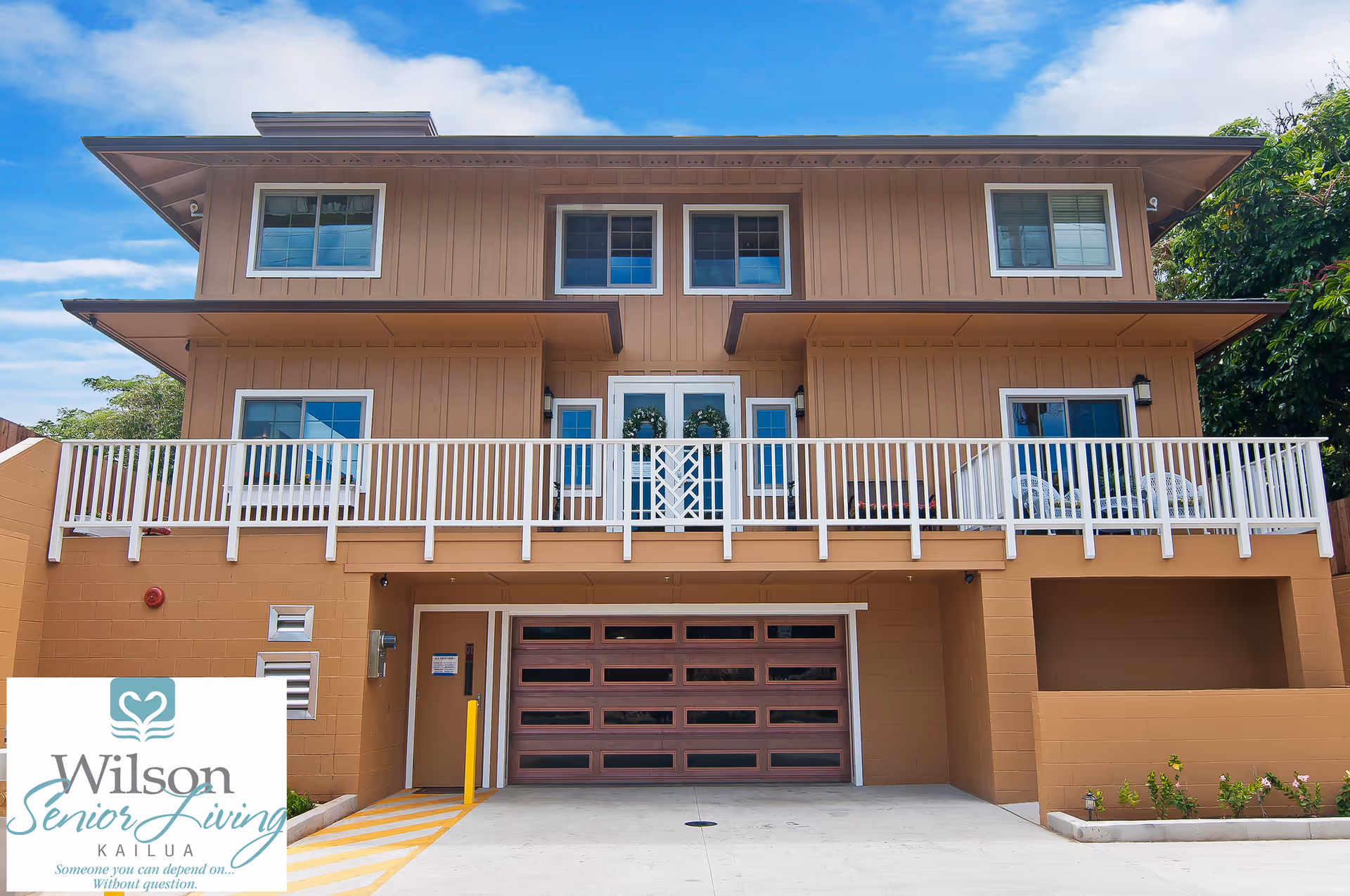 Exterior view of a two-story senior living facility building with a beige facade, white railings on the balcony, multiple windows, and a garage door on the ground level. There is a logo in the bottom left corner that reads 'Wilson Senior Living Kailua Someone you can depend on... Without question.'