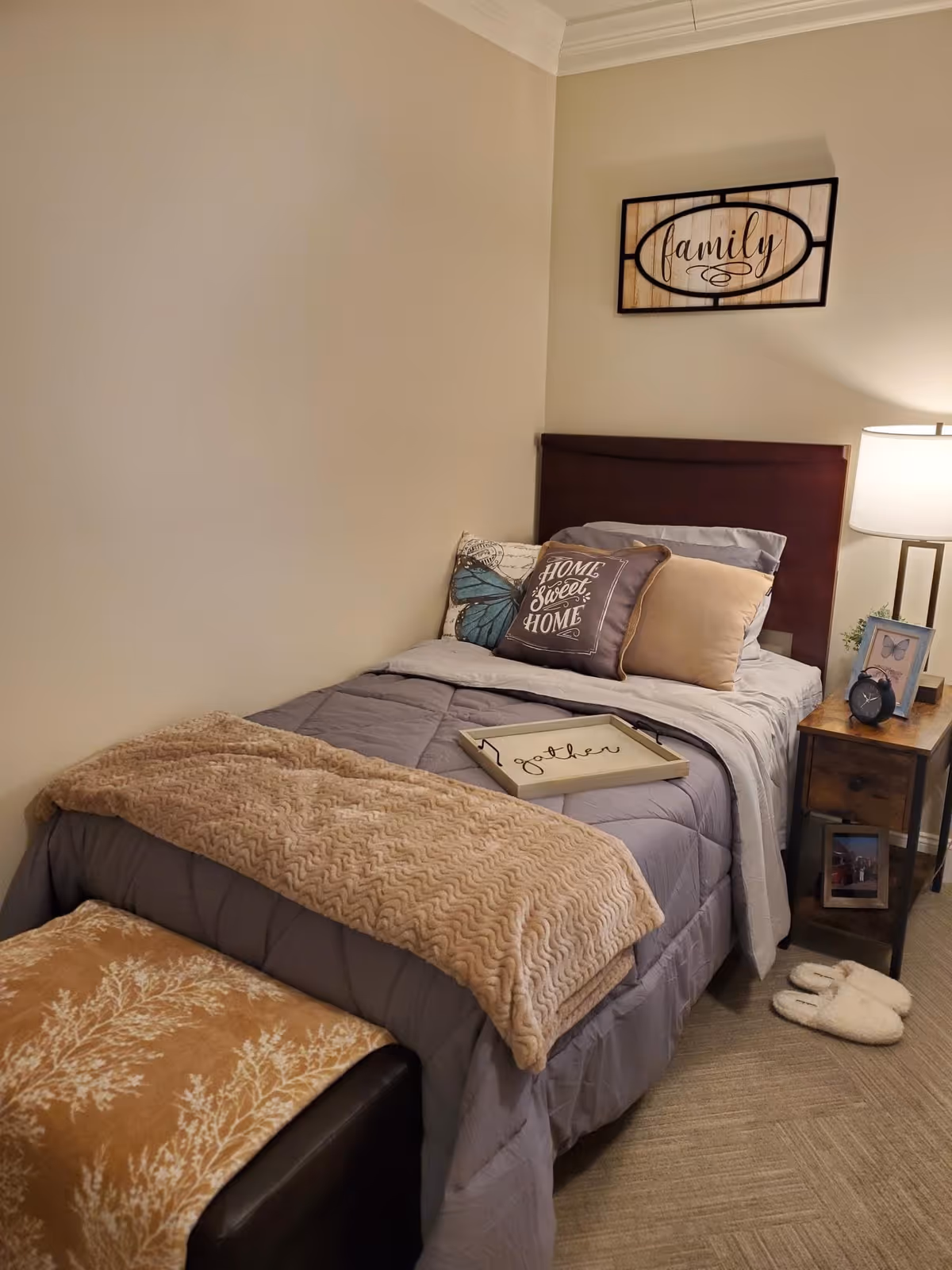 A cozy bedroom with a single bed made up with gray and beige bedding, decorative pillows including one that says 'Home Sweet Home,' and a beige throw blanket. A wooden nightstand with a lamp, a clock, a framed butterfly picture, and a small plant is beside the bed. A framed wall art with the word 'family' hangs above the headboard. A pair of white slippers is on the floor next to the nightstand.