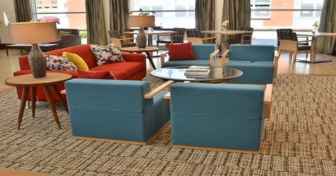 A spacious and well-lit living area with a red sofa, two blue armchairs, a round glass coffee table, and side tables with lamps. In the background, there are additional tables and chairs near large windows with curtains, allowing natural light to fill the room.