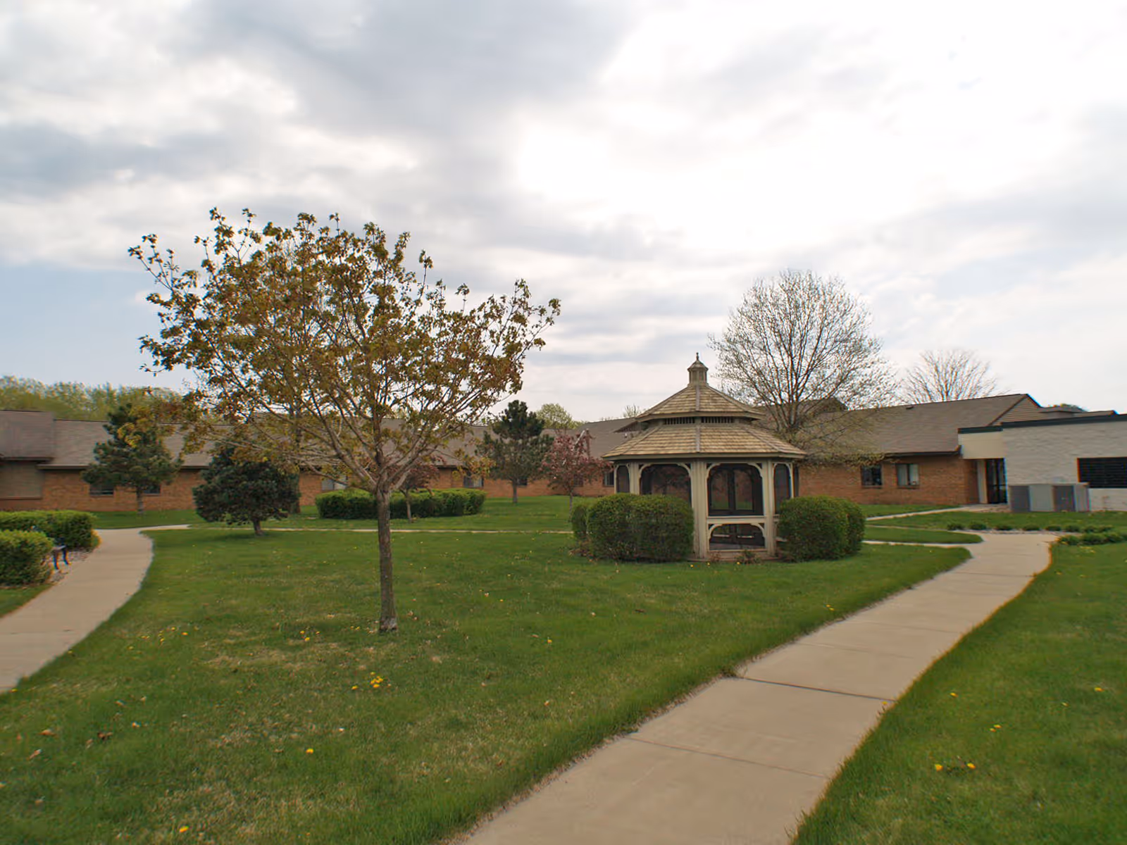 Courtyard with a central gazebo, lawns, trees, sidewalks, and a low brick senior living building in the background.