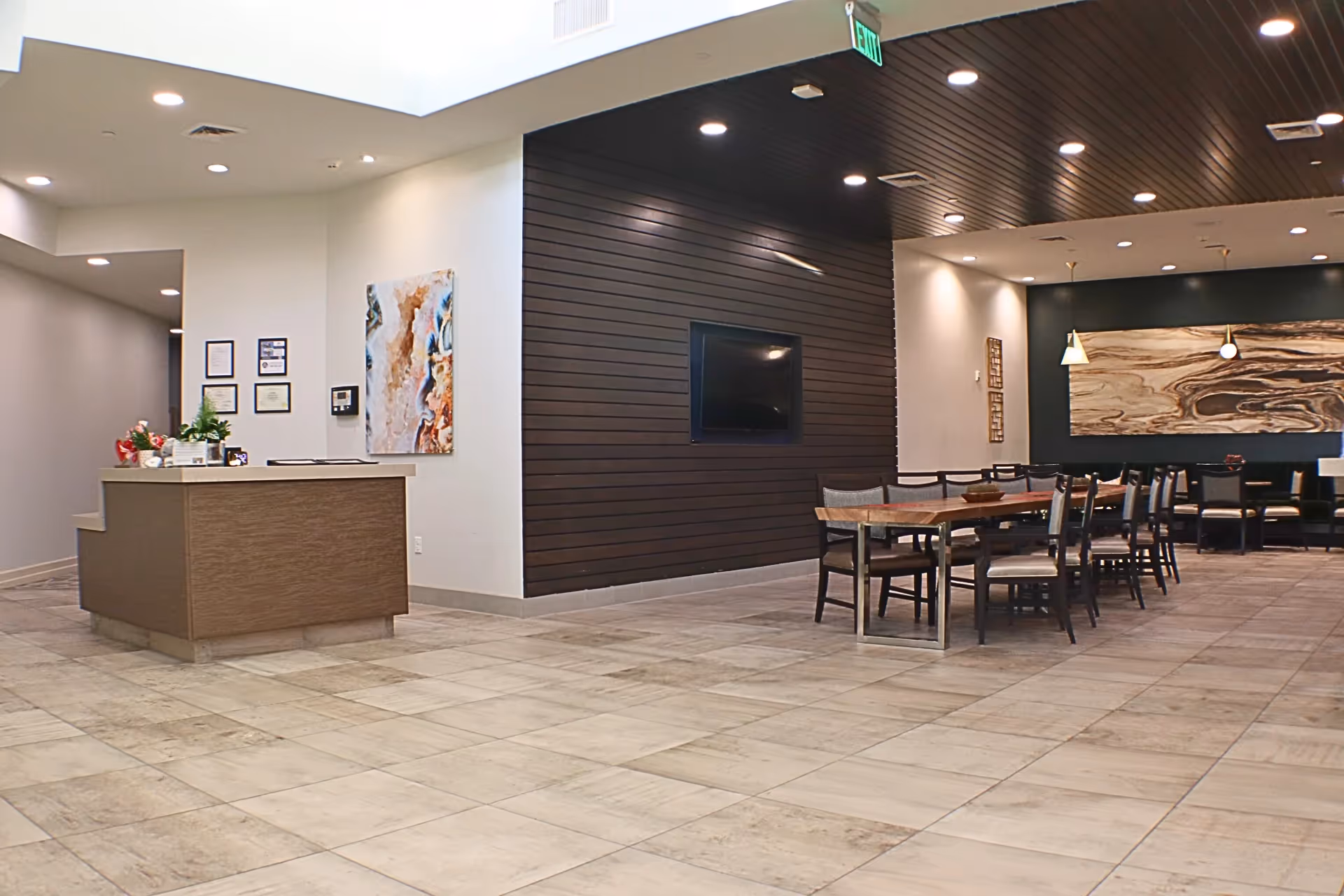 Interior view of a senior living facility common area with a reception desk on the left, a dark wood accent wall with a mounted TV in the center, and a dining area with tables and chairs on the right. The space has tiled flooring, recessed lighting, and modern decor including wall art and plants.