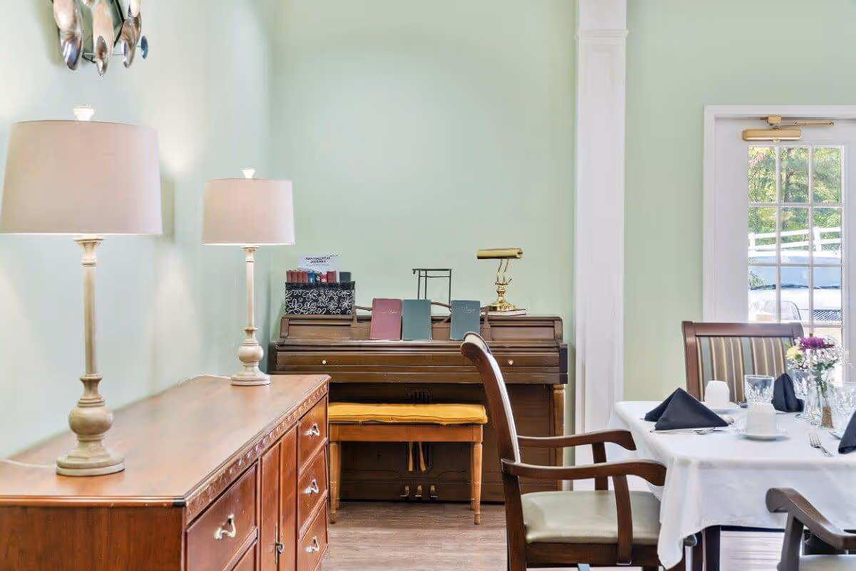 A cozy interior room with light green walls featuring a wooden sideboard with two table lamps, a piano with sheet music and a brass lamp on top, and a dining table set with white tablecloth, black napkins, glassware, and a small flower arrangement. A glass door with a view of greenery outside is visible in the background.