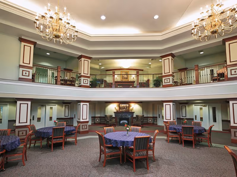 Spacious interior common area with round tables covered in dark blue tablecloths and wooden chairs arranged around them. The room features a two-story design with a balcony railing on the upper level, decorative columns with wood trim, and two large chandeliers hanging from the ceiling. There is a fireplace with two armchairs and artwork above it on the far wall, and the overall decor is elegant and traditional.