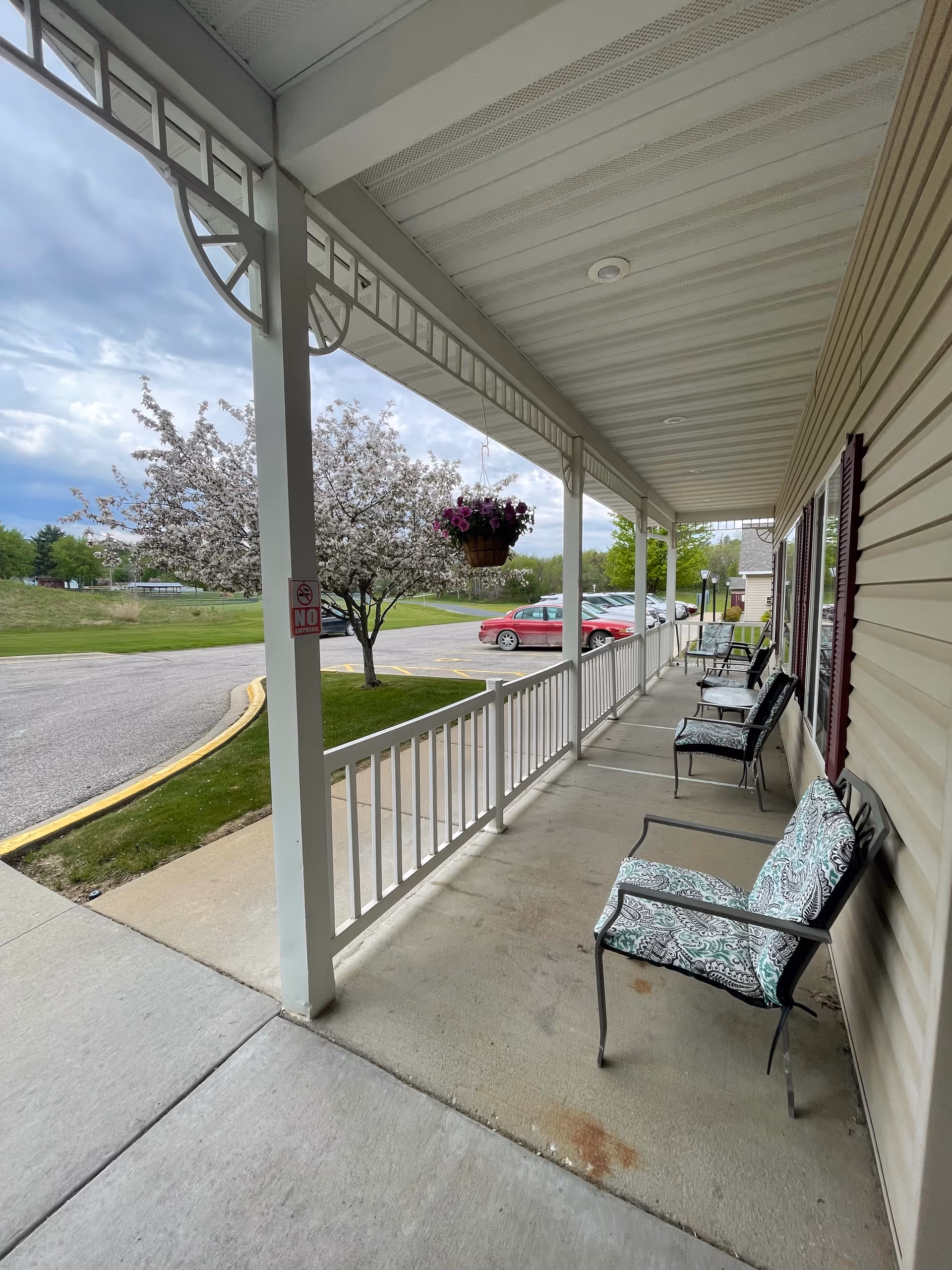 Covered outdoor porch area with several cushioned metal chairs lined up against the building wall. A hanging basket with purple flowers is suspended from the porch ceiling. In the background, there is a parking lot with several cars and a tree with white blossoms. The sky is partly cloudy.