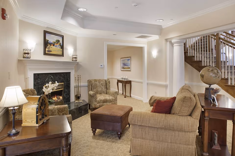 A cozy living room with beige walls and carpet, featuring a fireplace with a black marble surround and a painting above it. The room has a patterned armchair, a brown ottoman, and a beige sofa with a red pillow. There are two wooden side tables, one with a lamp and a decorative item, and the other with a globe and small framed pictures. A staircase with wooden steps and white balusters is visible in the background.