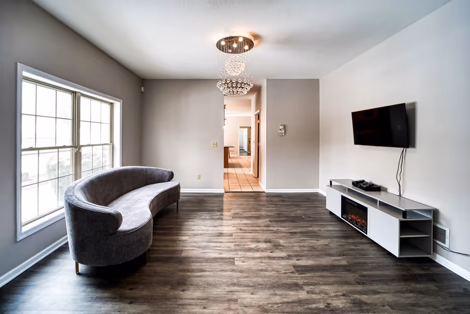 A modern living room with a curved gray sofa positioned near a large window on the left. The room features dark wood flooring, a wall-mounted flat-screen TV on the right, and a white media console with an electric fireplace below the TV. A decorative chandelier hangs from the ceiling, and a hallway with tiled flooring is visible in the background.