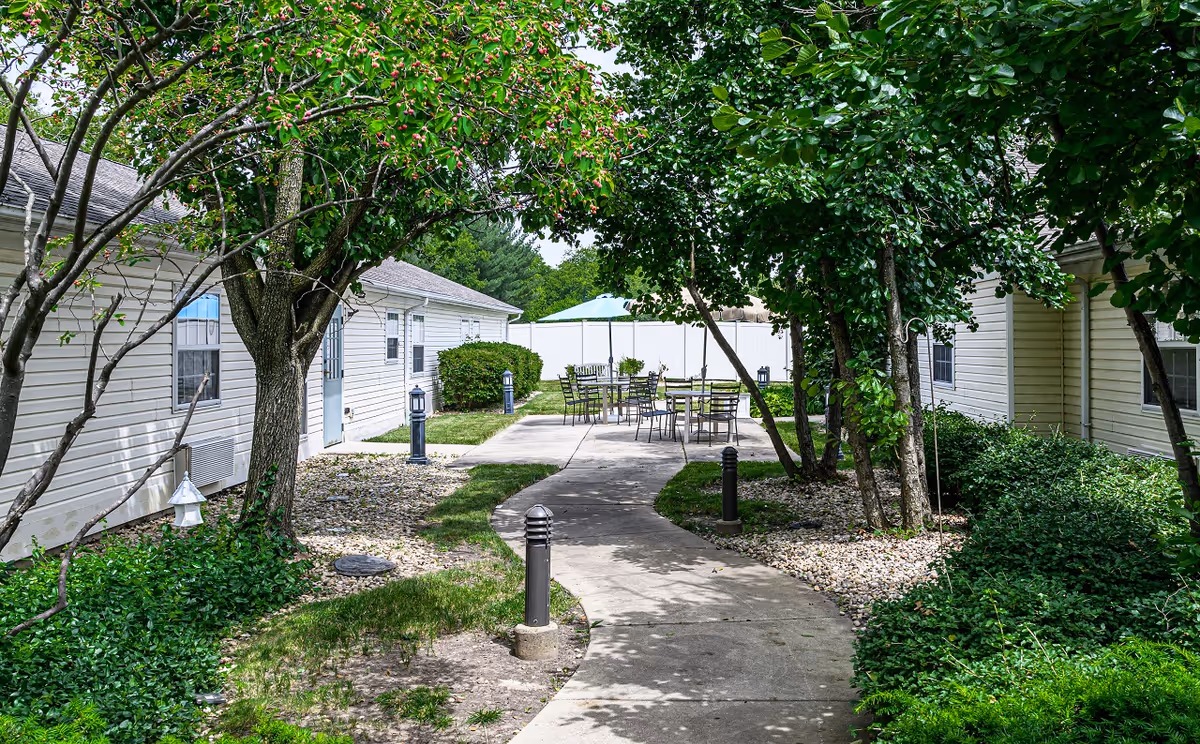 Outdoor courtyard area with a curved concrete pathway leading to a seating area with metal tables and chairs under a blue umbrella. The courtyard is surrounded by green trees, bushes, and two single-story beige buildings with windows and doors.