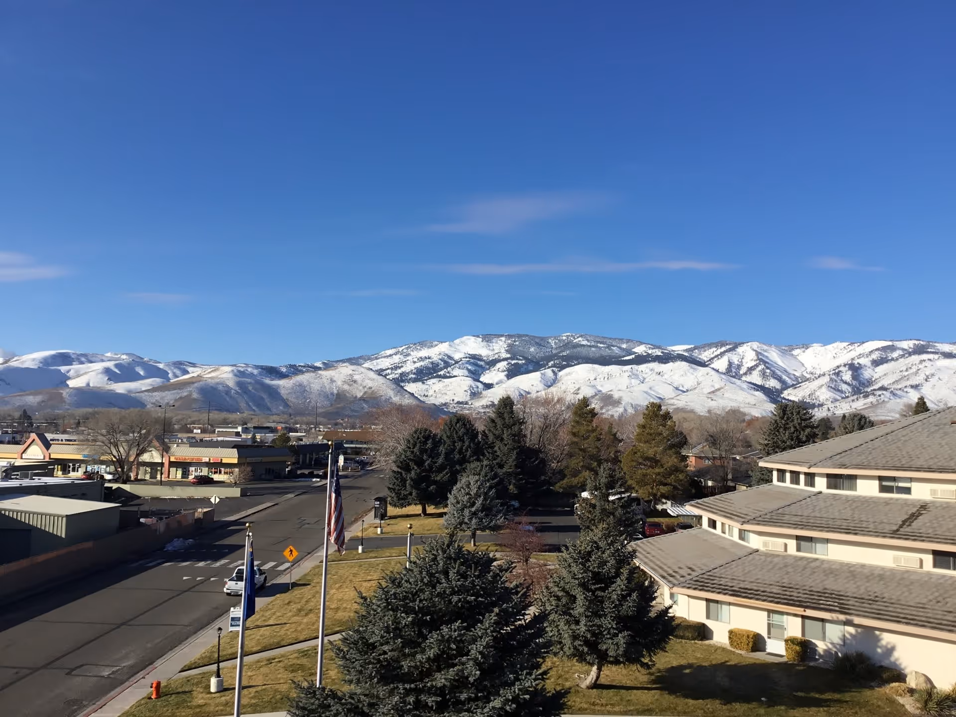 View of a street with buildings and trees in the foreground, with snow-covered mountains under a clear blue sky in the background.