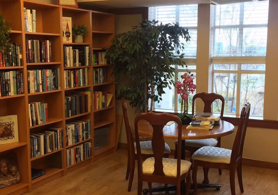 A cozy reading area with wooden bookshelves filled with books on the left and a round wooden table with four cushioned chairs in front of a large window. A potted plant and a vase with pink flowers are on the table, and natural light comes through the window.