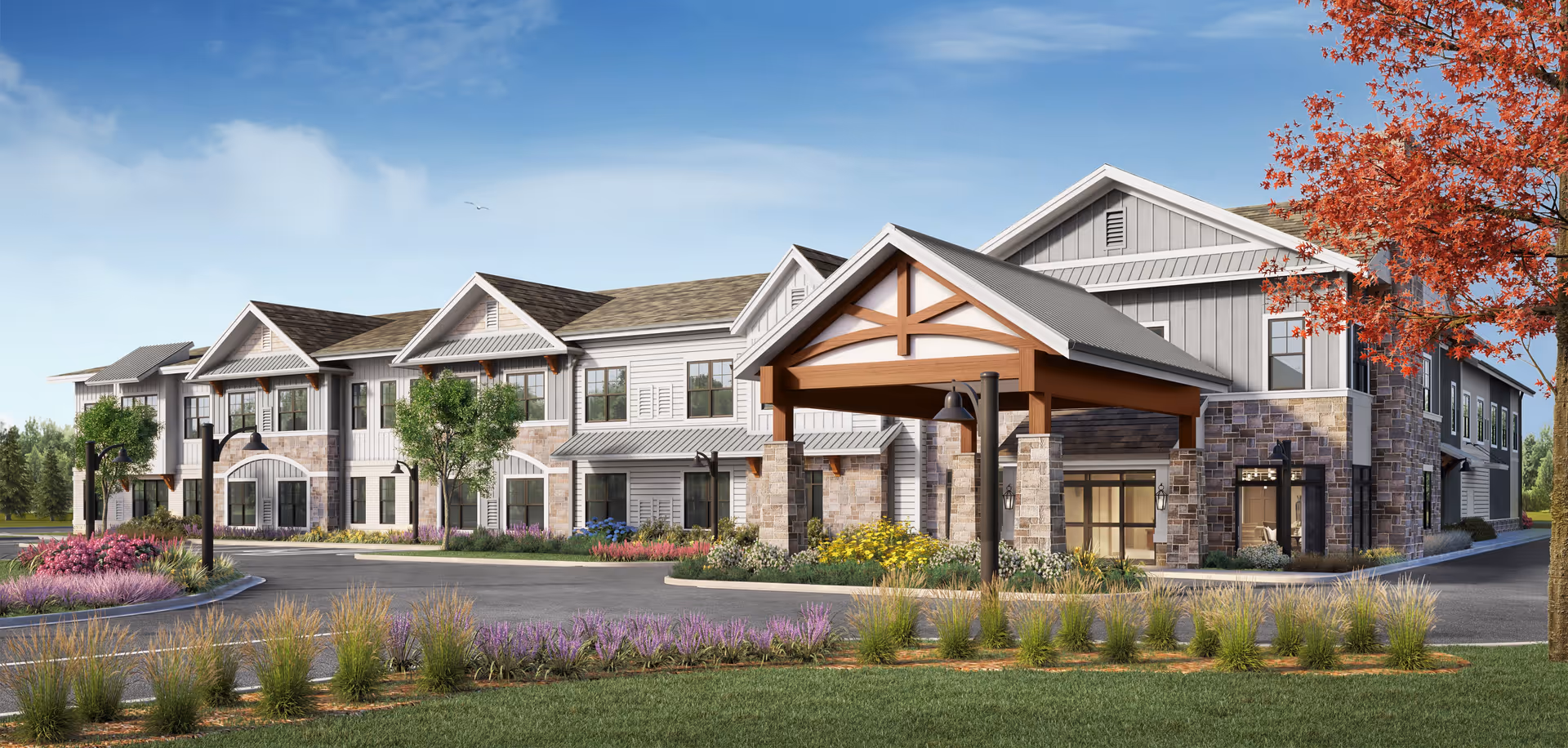 Exterior view of a two-story senior living facility building with a covered entrance supported by stone pillars. The building features a combination of stone and siding with multiple windows. There is a landscaped area with various plants, flowers, and trees, including one with red leaves, under a clear blue sky.