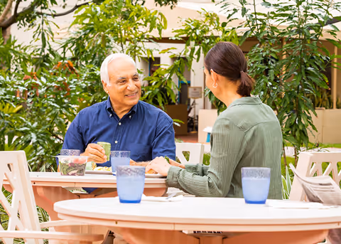 An elderly man and a woman sitting at a round outdoor table having a conversation and enjoying a meal, surrounded by green plants in a garden-like setting.