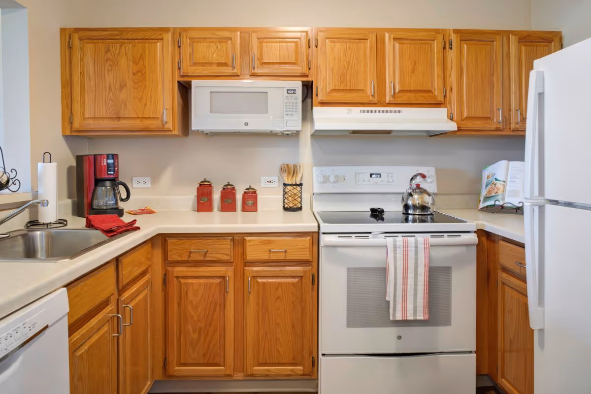 Bright kitchen with oak cabinets, white stove and microwave, a kettle on the stove, coffee maker by the sink, and a refrigerator at right.