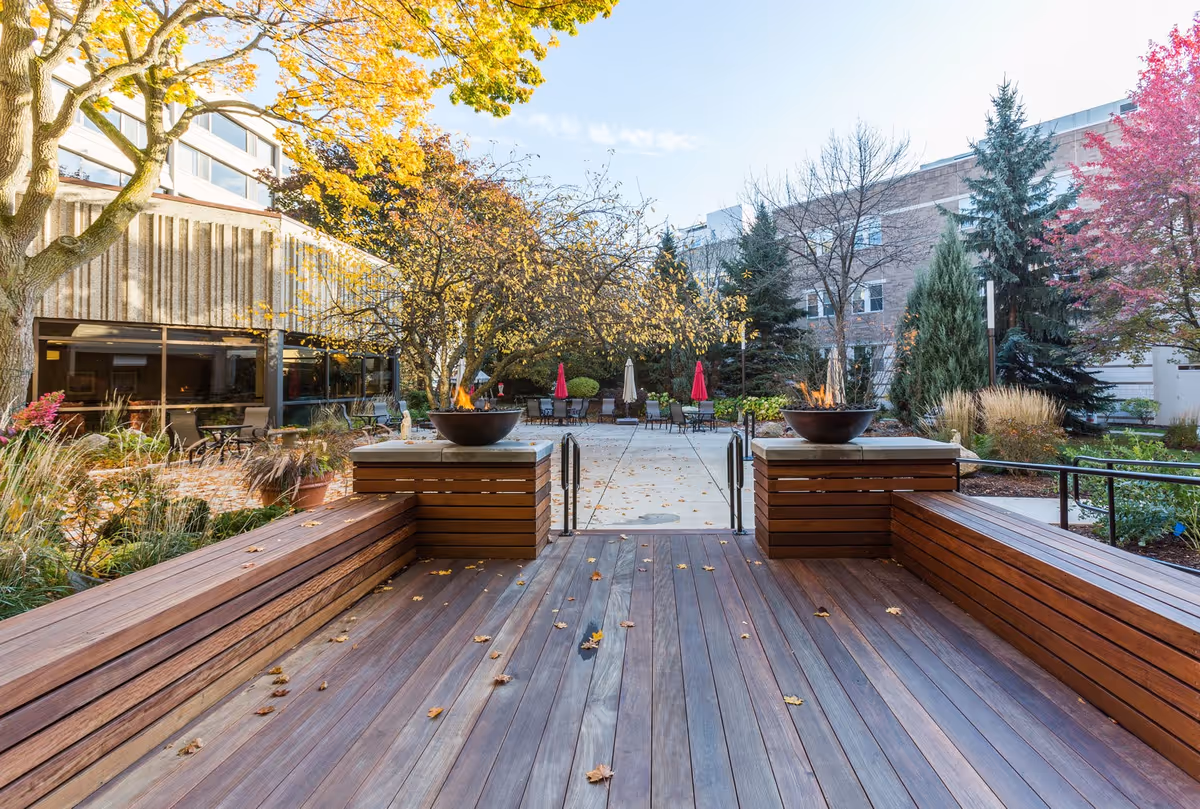 Outdoor patio area with wooden benches and two fire pits on raised platforms. The patio is surrounded by trees with autumn foliage and some outdoor tables with umbrellas in the background. A multi-story building is visible behind the trees.