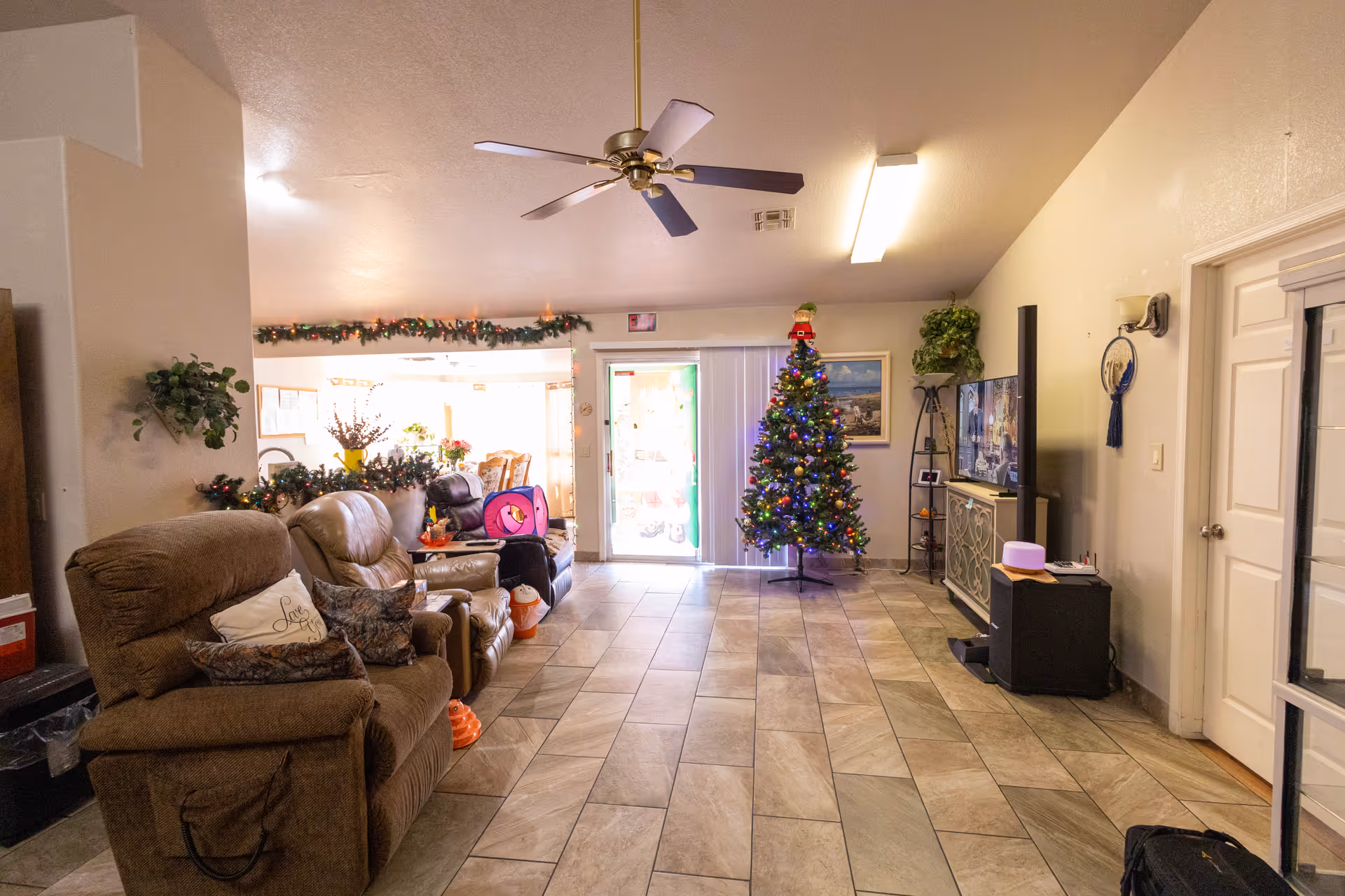 Spacious living room with recliner sofas, a decorated Christmas tree, TV and tiled floor leading to a sliding glass door.