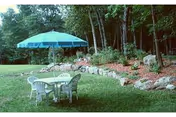 Plastic outdoor table and chairs with a blue umbrella on a lawn beside a landscaped rock border and trees.