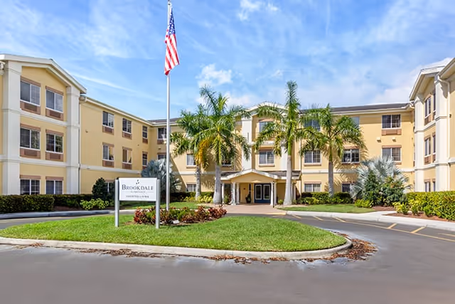 Front exterior view of Brookdale Palmer Ranch, a three-story senior living facility with beige and white walls, palm trees, an American flag on a flagpole, and a circular driveway with landscaped greenery.