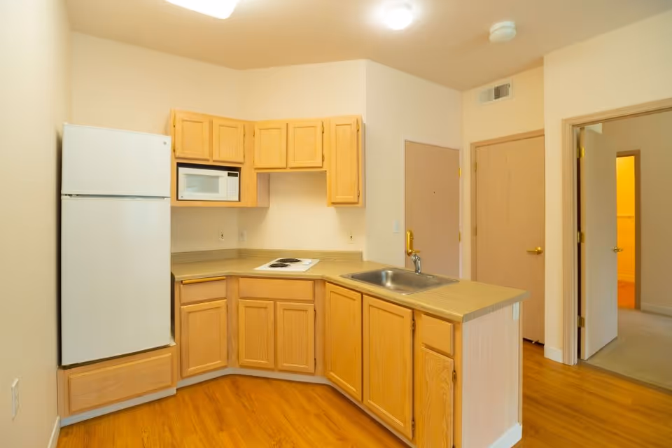 Small kitchen with light wood cabinets, a white refrigerator and microwave, a sink in the peninsula, and hardwood floors.