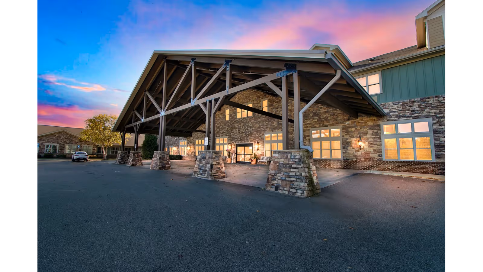 Exterior view of The Phoenix at Lake Lanier facility during sunset, showing a large covered entrance supported by stone and wooden beams, with warm lights glowing from the windows and a car parked to the left.