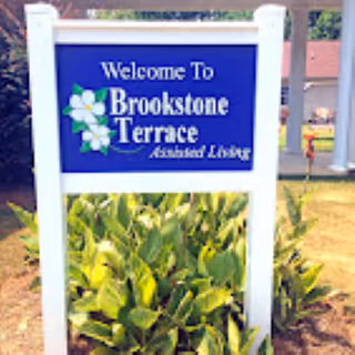 A blue and white sign that reads 'Welcome To Brookstone Terrace Assisted Living' with a white flower graphic, surrounded by green plants and grass in an outdoor setting.
