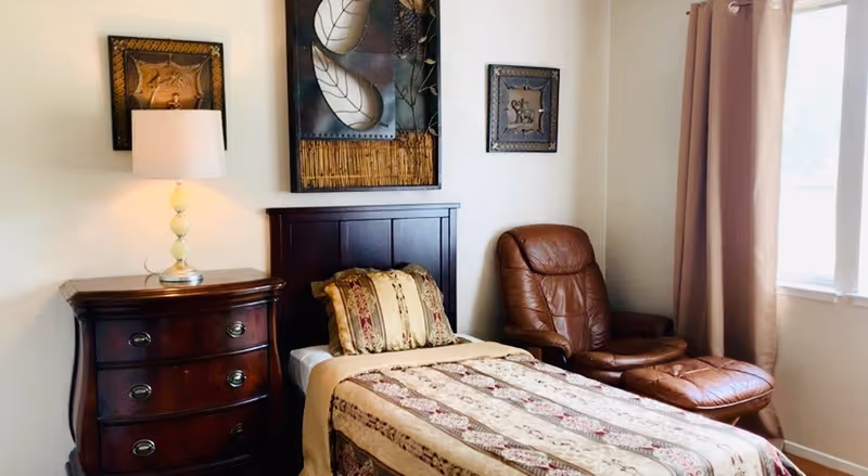 A cozy bedroom with a single bed covered in a patterned beige and brown bedspread and matching pillows. Next to the bed is a dark wooden nightstand with three drawers and a lamp with a white shade. On the wall above the bed are three decorative framed artworks. To the right of the bed is a brown leather armchair with a matching ottoman, positioned near a window with beige curtains.