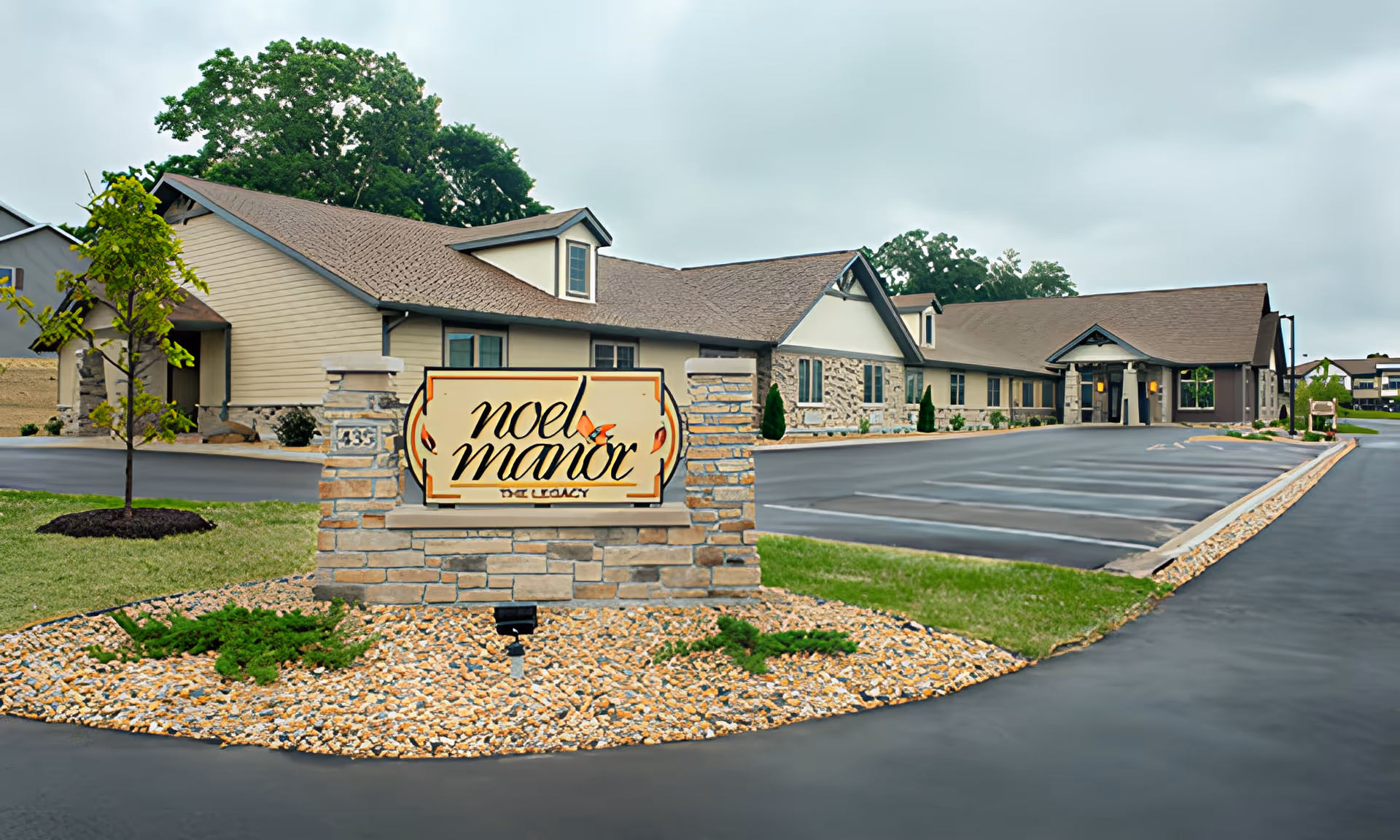 Exterior view of The Legacy at Noel Manor building with a stone sign in front displaying the facility name. The building has a beige and stone facade with a brown roof, surrounded by a paved parking lot and landscaped areas with small trees and shrubs.