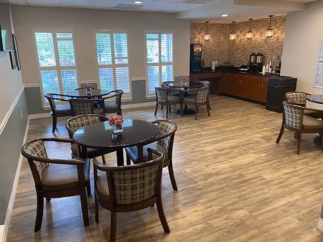 A bright dining area with several round tables and cushioned chairs arranged around them. The room has large windows with white shutters allowing natural light to enter. In the back corner, there is a counter with a coffee machine, cups, and other beverage supplies. The floor is covered with light wood-style flooring, and the walls are painted in neutral tones.
