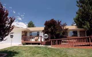 Single-story residential building with a wooden ramp and porch, surrounded by green grass, trees, and a clear blue sky.