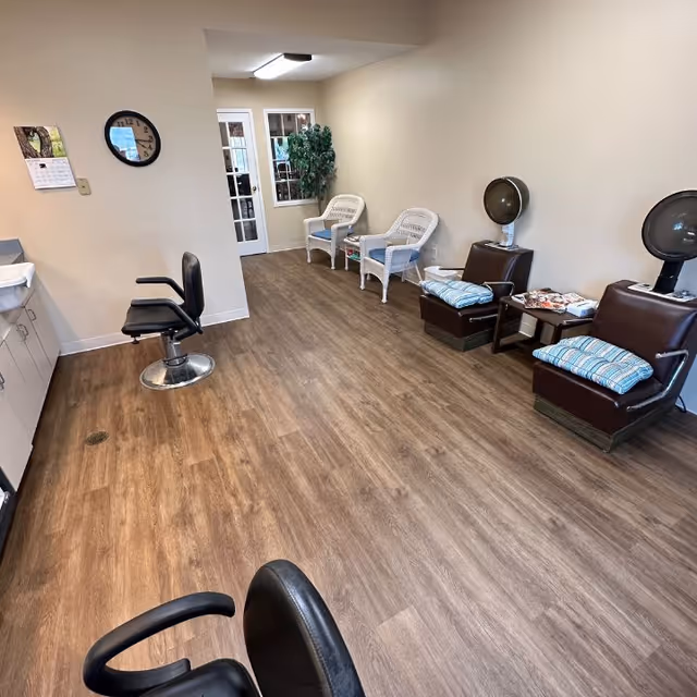 Interior view of a salon area in Seminary Estates featuring two black salon chairs, two brown hair dryer chairs with blue striped cushions, two white wicker chairs, a small table with magazines, a wall clock, a calendar, and a potted plant near a glass door.
