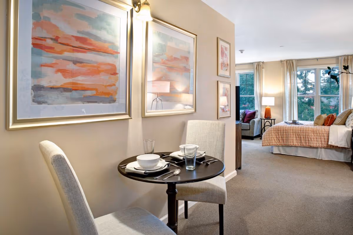 A cozy interior space featuring a small round dining table set for two with white bowls, glasses, and silverware. Two upholstered chairs are positioned at the table. The wall behind the table has two large abstract paintings with warm tones. In the background, there is a bedroom area with a bed covered in a patterned orange bedspread, multiple pillows, a bedside table with a lamp, and large windows with beige curtains letting in natural light.